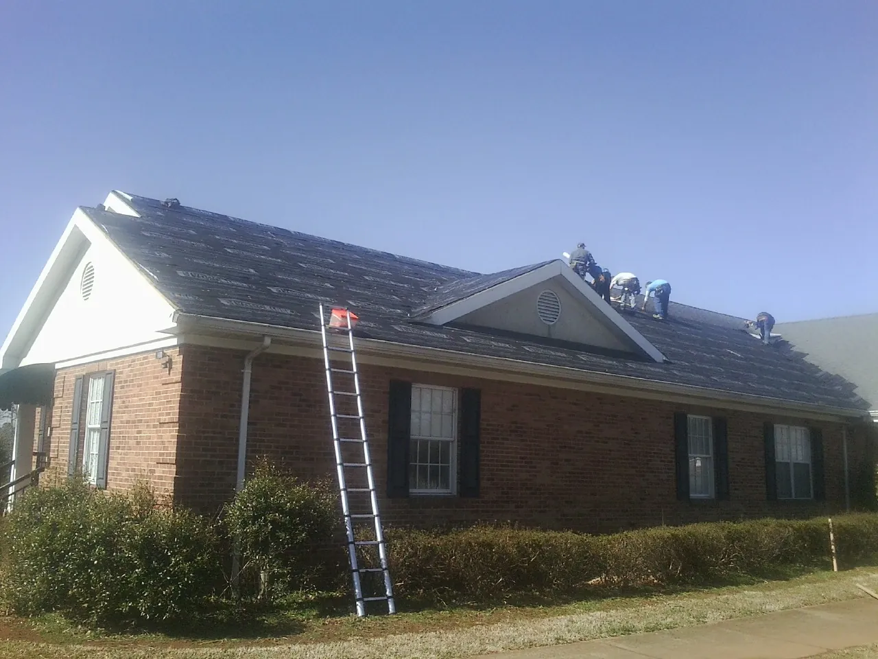 Crew tearing shingles off a brick ranch roof with a ladder set — Emergency Roof Repair work by our crew in Pelzer