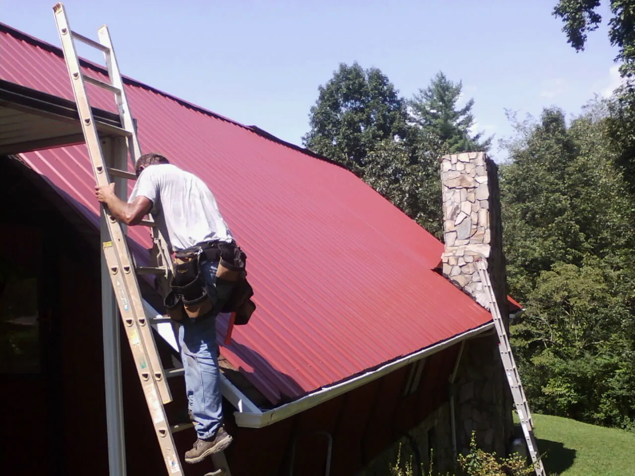 Roofer on a ladder beside a red metal roof and stone chimney — Soffit & Fascia Repair by Roofing Brothers in Pelzer